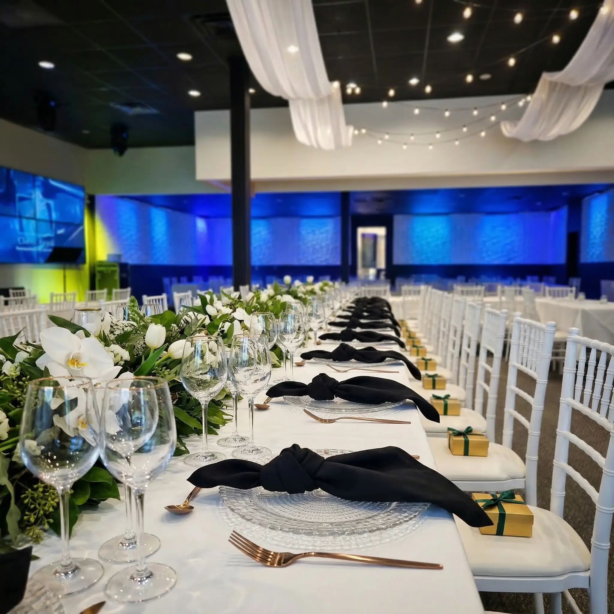 Elegant dining setup with white chairs, floral centerpieces, and neatly arranged tableware under soft lighting.