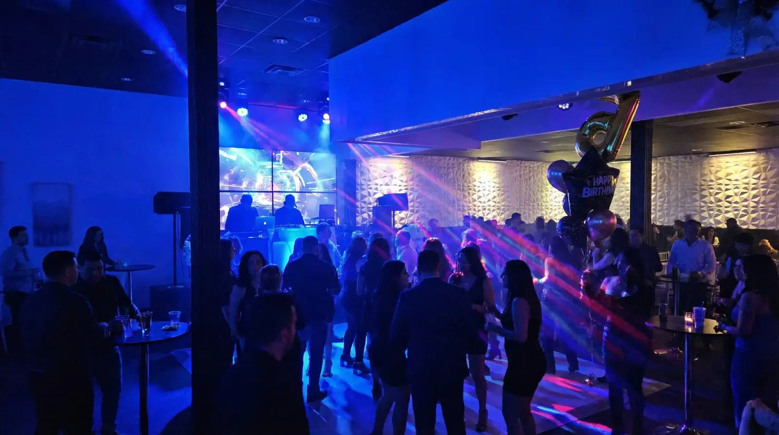 Crowd dancing in a vibrant nightclub with colorful lights and a DJ setup in the background.